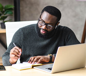 Smiling African-American guy sits at the desk in modern office, young black man in glasses is watching at laptop screen and writing notes in notebook