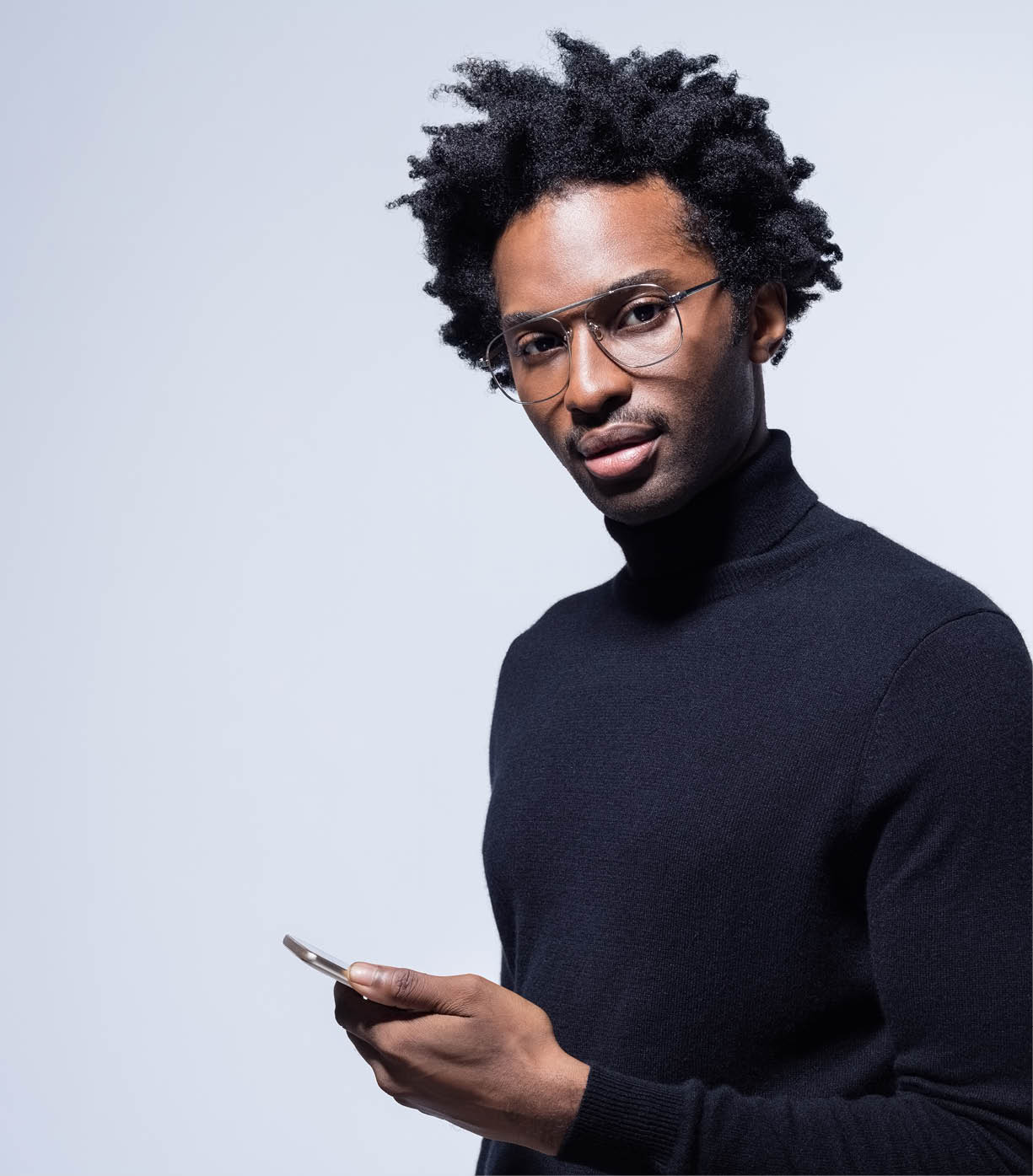 Afro american young man wearing black turtleneck and glasses holding smart phone in hand, looking at camera  Studio shot on grey background 